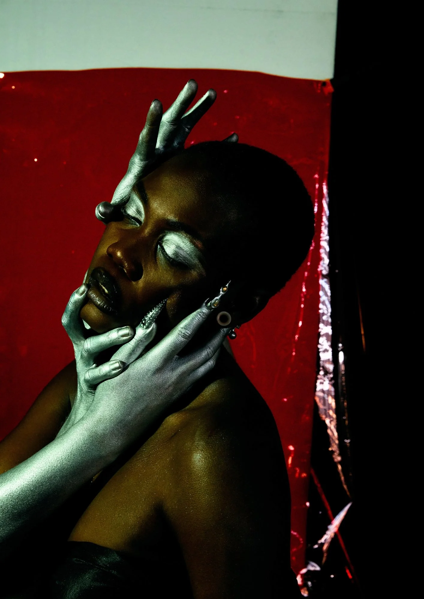A woman with short hair and dark skin, partially covered in metallic silver paint, is posing with her eyes closed and her fingers touching her face. She has artistic makeup with silver and dark shades, and the background features red and black elemen