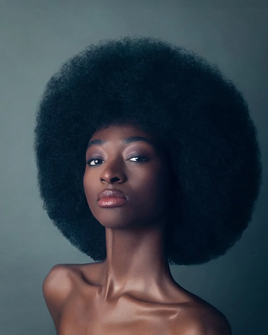 Portrait of a Black woman with natural, voluminous afro hairstyle, looking confidently at the camera with smooth skin and subtle makeup against a neutral gray background.