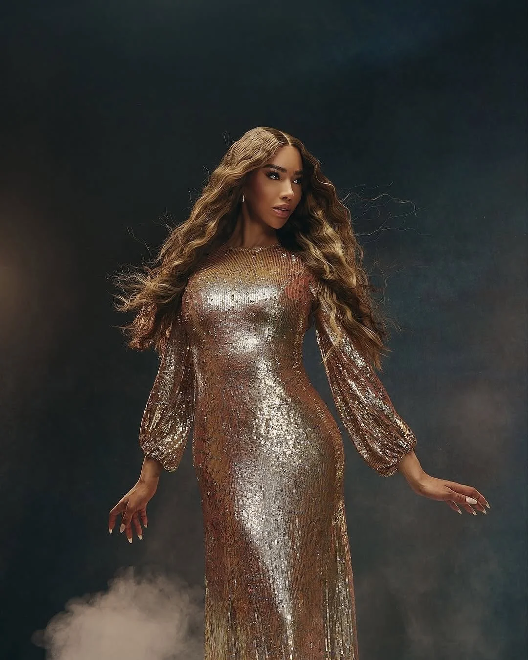 A woman with long, wavy hair wearing a sparkling, gold, long-sleeved gown standing against a dark background with some fog at the bottom.