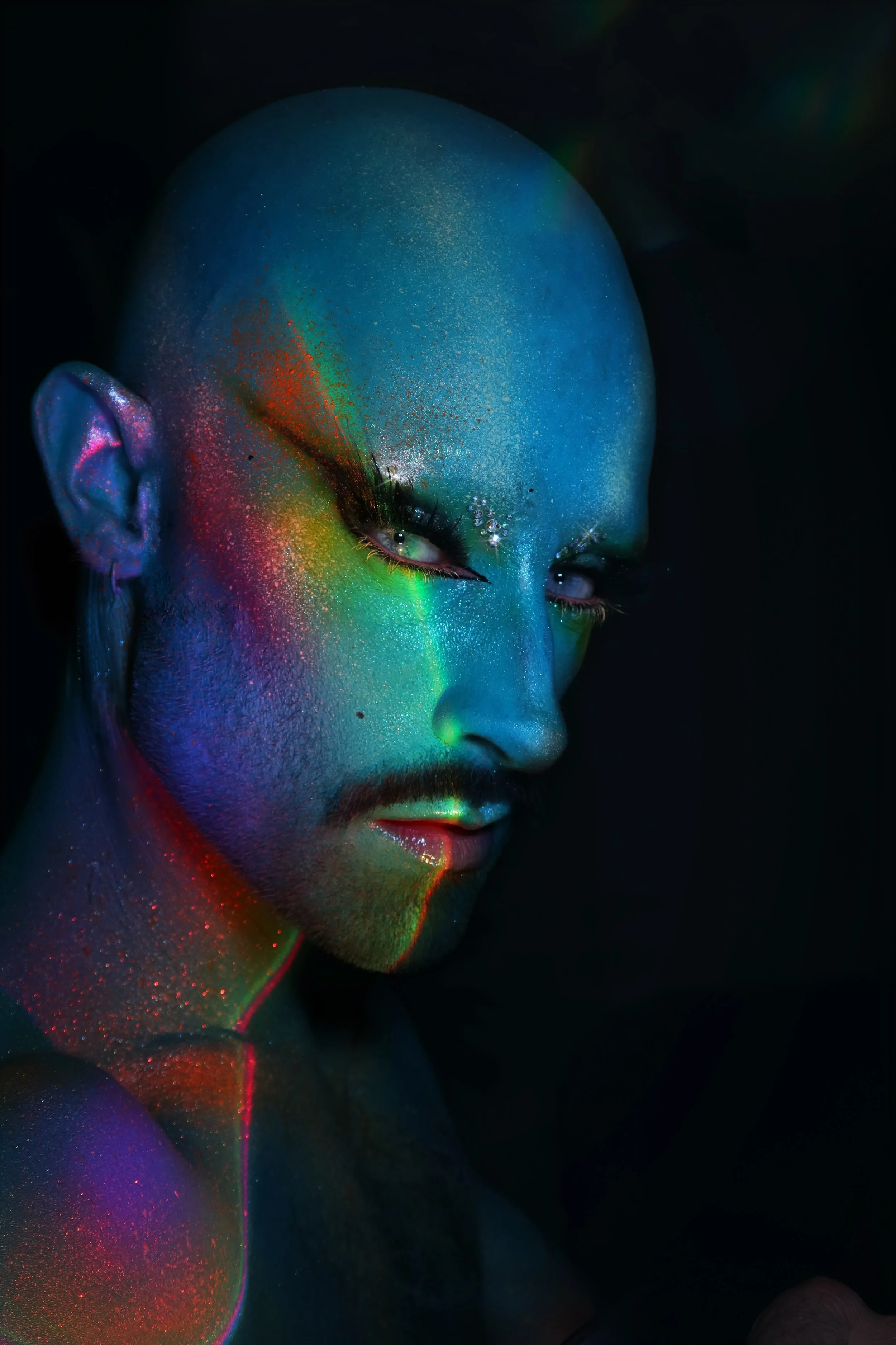 A person with vibrant, luminous facial makeup featuring a mix of neon colors such as blue, green, yellow, red, and purple, against a pitch-black background. The makeup includes glitter and highlights around the eyes and face.