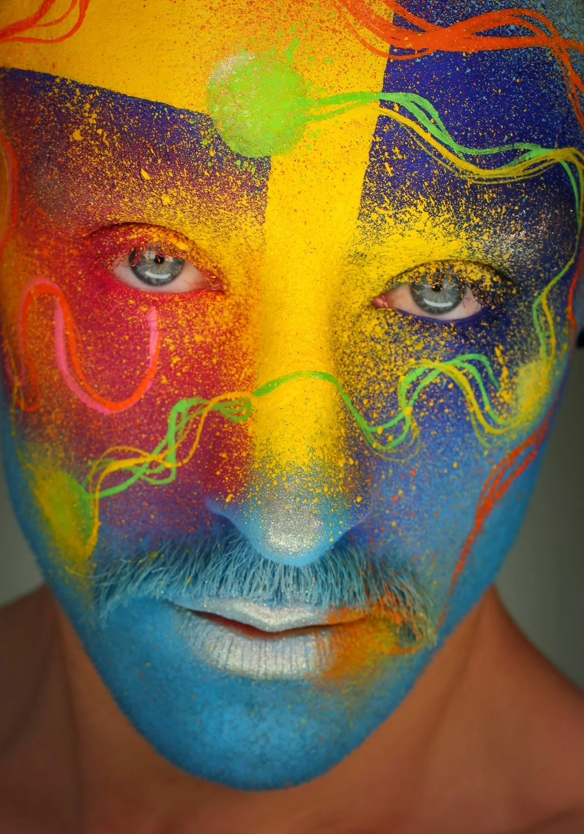 Close-up of a person's face painted with vibrant colors and intricate designs, with the person's natural eyes visible through the face paint.