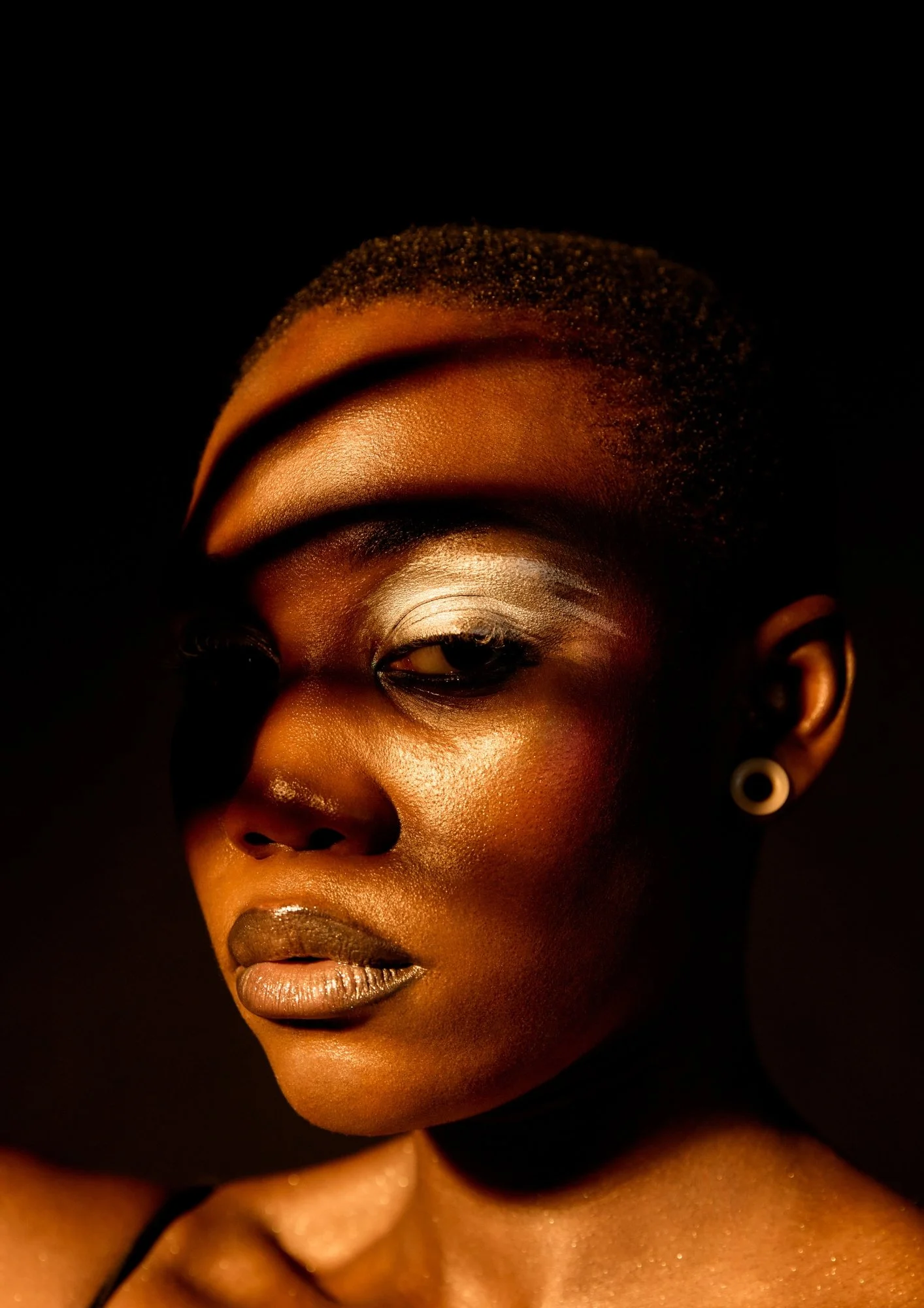Close-up of a woman with dark skin, gold makeup, and earrings against a black background.