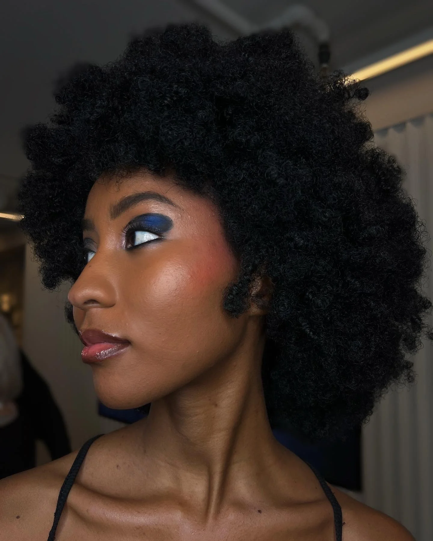 Close-up of a woman with dark curly hair, wearing blue eyeshadow, with natural makeup, wearing a black spaghetti strap top.