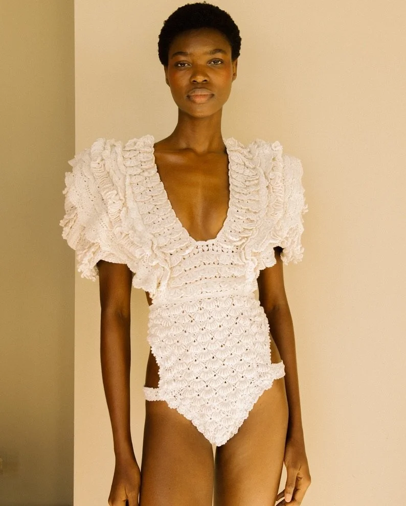 A woman with dark skin and short curly hair posing indoors against a beige wall, wearing a white crocheted outfit with ruffled details and cutouts.