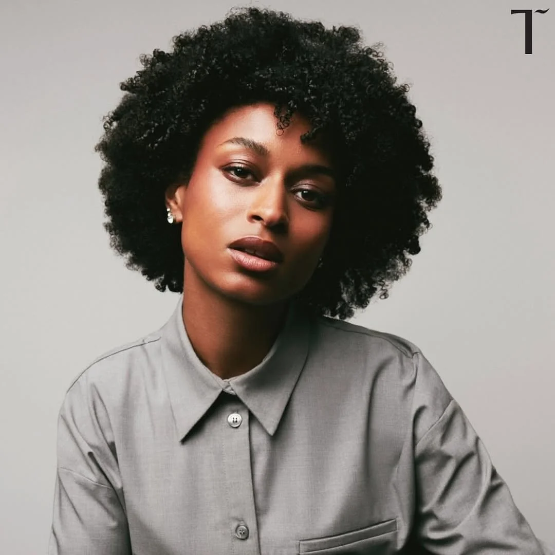 A woman with dark skin and curly black hair wearing earrings and a gray button-up shirt, looking at the camera against a plain background.