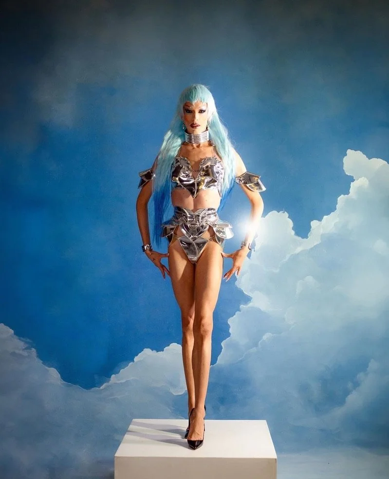A woman with long, light blue hair dressed in a futuristic, metallic outfit standing on a white platform against a backdrop of blue sky with clouds.