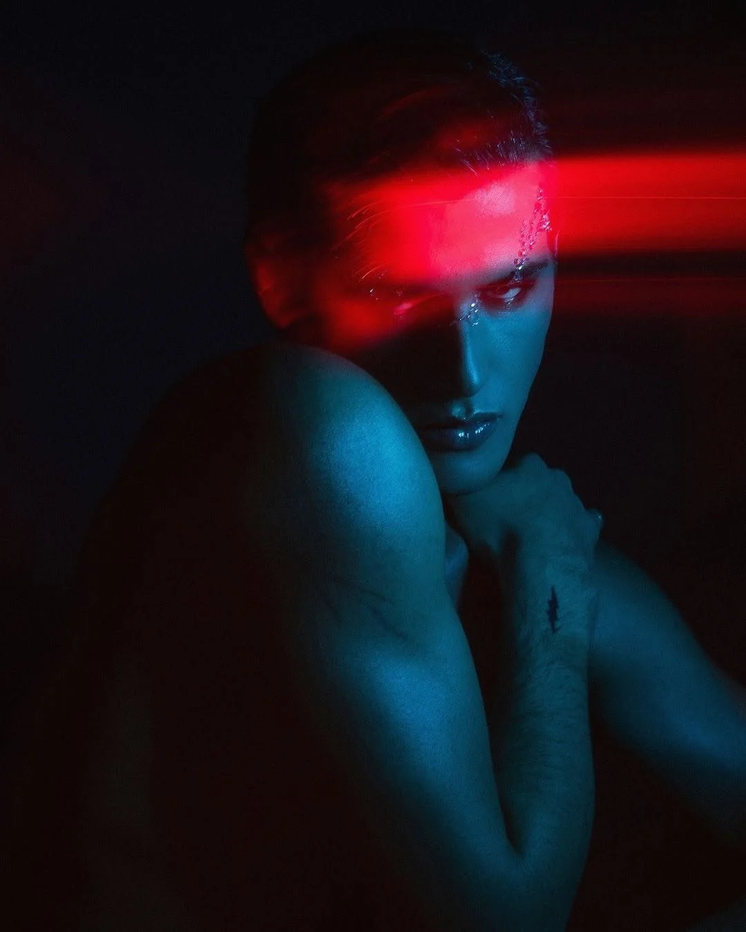 A woman with slicked-back hair, illuminated by red and blue lighting, gazes intensely at the camera with her chin resting on her shoulder, in a dark setting.
