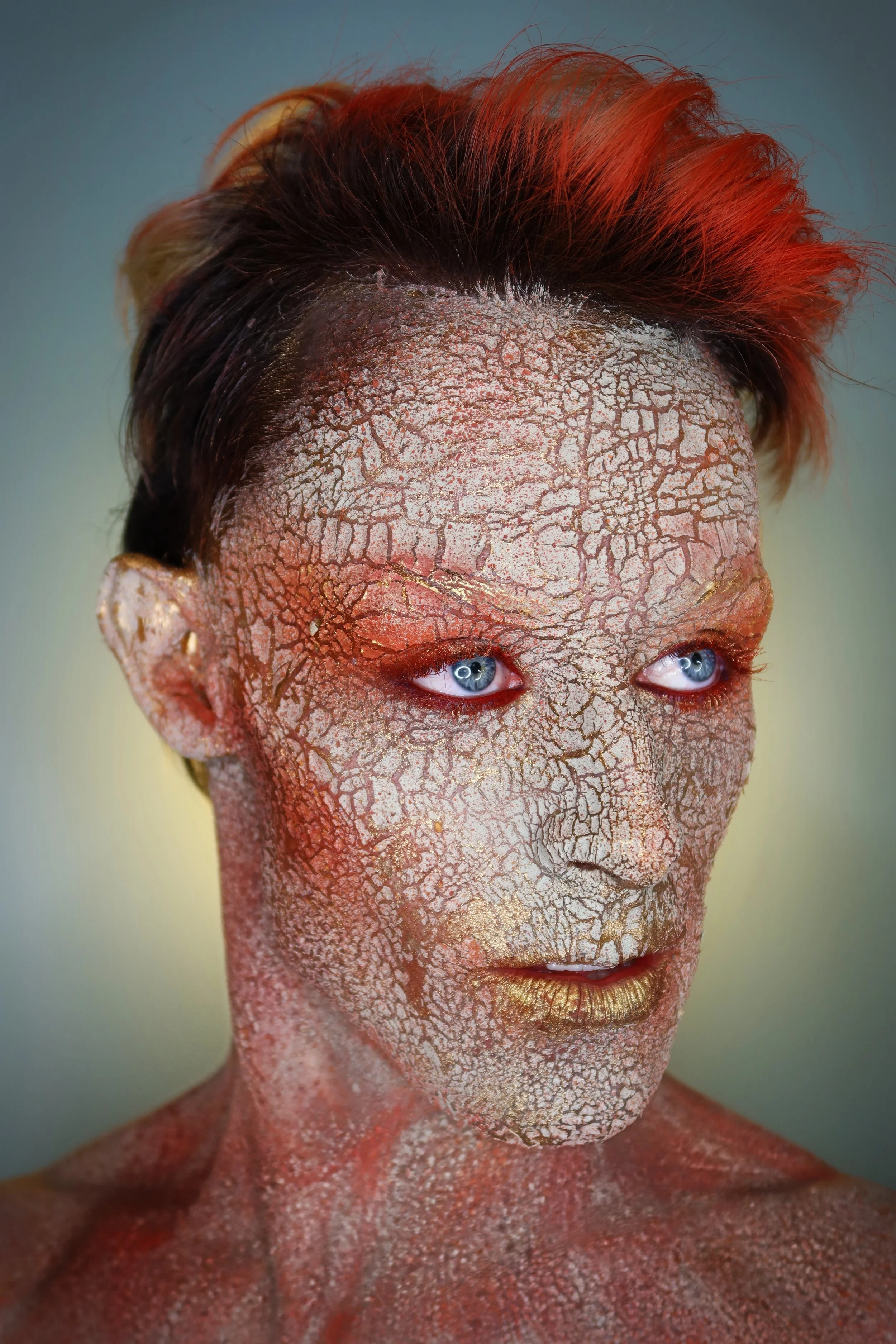 A person with bright red hair and striking blue eyes, their face, neck, and shoulders are covered in cracked, metallic-looking paint in shades of gold, red, and white.