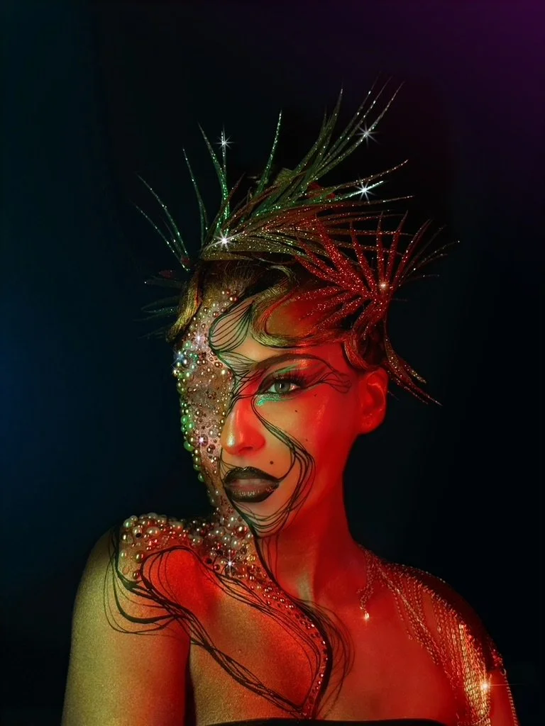 A woman with elaborate headpieces, glitter, pearls, and artistic makeup illuminated with red and green lighting at a dark background.