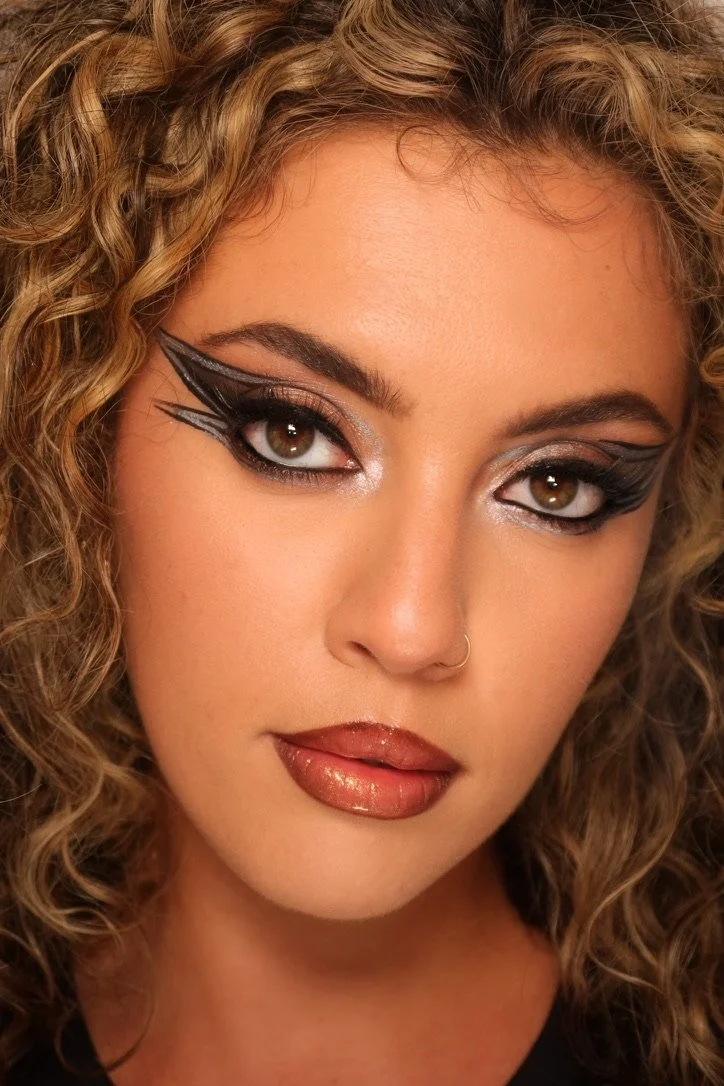 Close-up of a woman with curly blonde hair wearing bold black eyeliner and metallic eyeshadow, with glossy lips and a small nose ring.