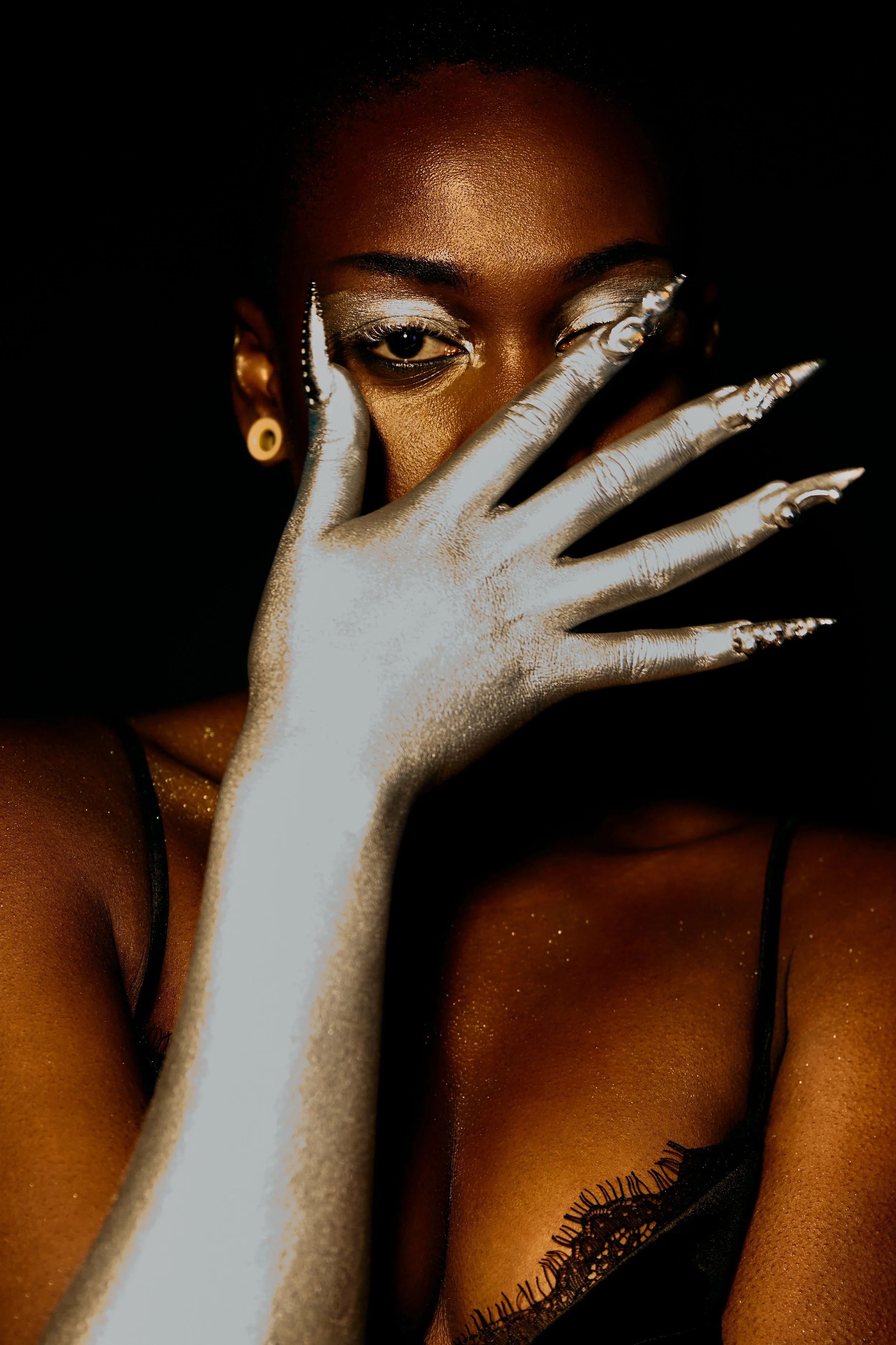 Close-up of a woman with dark skin, gold eye makeup, black lace lingerie, and long painted nails, covering her face with her hand against a black background.