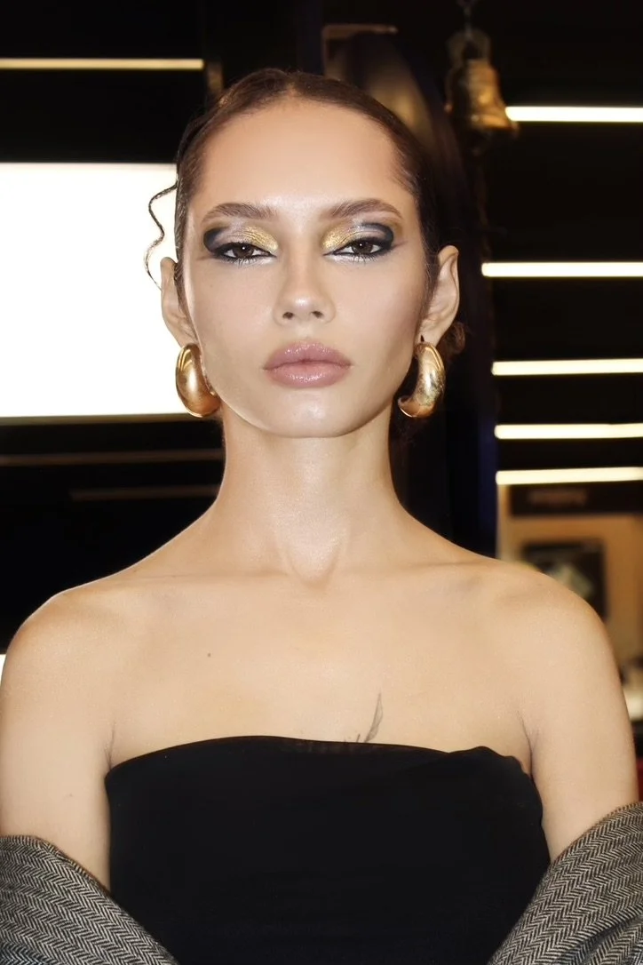 A woman with styled brown hair, bold eye makeup with gold and black eyeshadow, nude lipstick, wearing large gold earrings, a black off-the-shoulder top, and a patterned jacket draped over her arms, standing indoors with modern lighting and decor in t