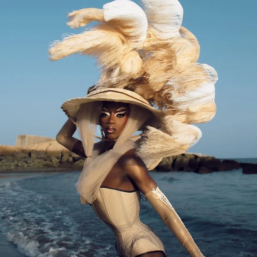 A woman standing on a beach with an elaborate large feathered headdress and a form-fitting beige dress.