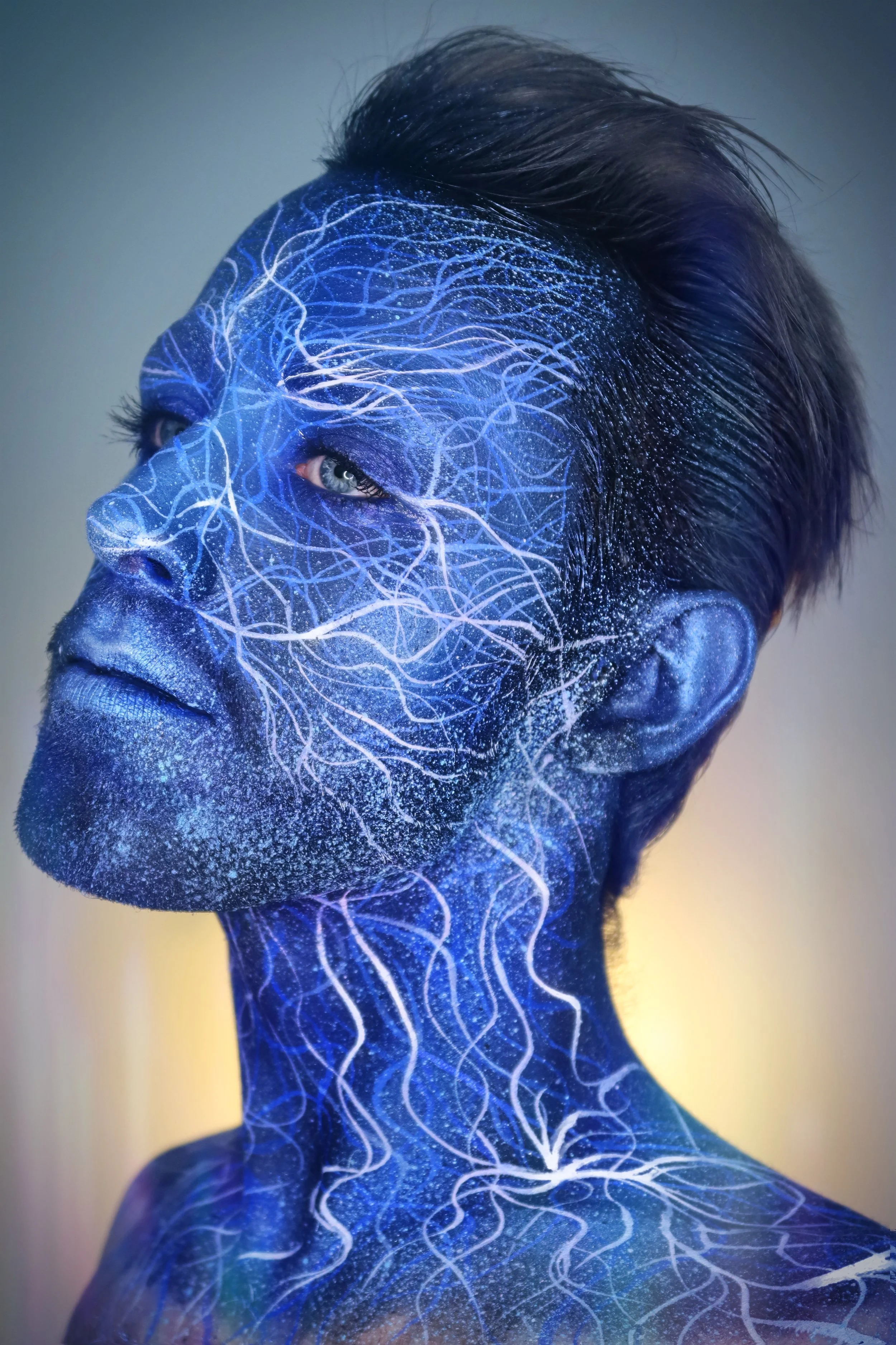 A person with blue painted skin and white neural network-like patterns covering their face and neck, with dark hair styled upward.