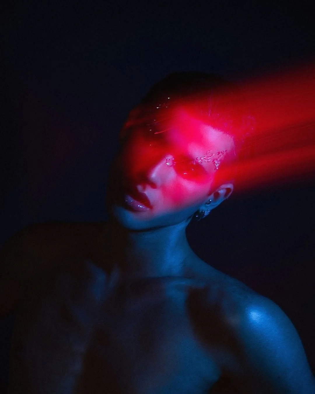 A person with short hair and earrings is illuminated by red and blue lights in a dark setting, creating a futuristic or artistic effect.