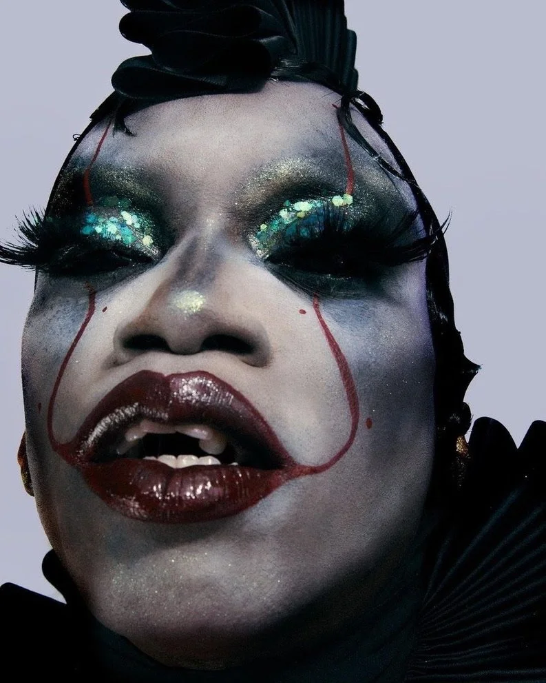 Close-up of a person's face with dramatic makeup, including glittery eyeshadow, long false eyelashes, dark red lipstick, and face paint resembling a clown or theatrical character.