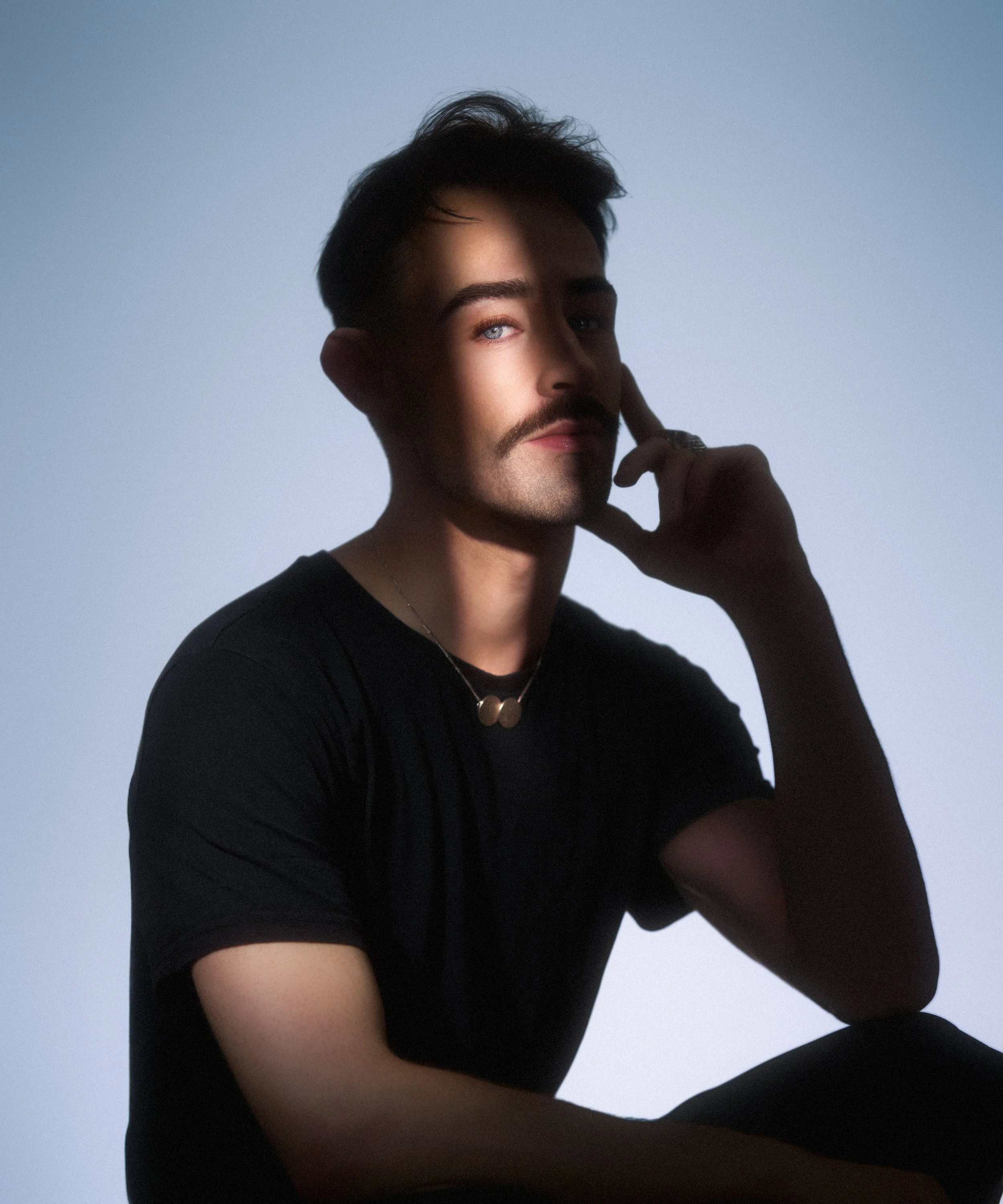 Portrait of a young man with dark hair and blue eyes, wearing a black shirt and a silver ring, sitting against a plain light blue background.