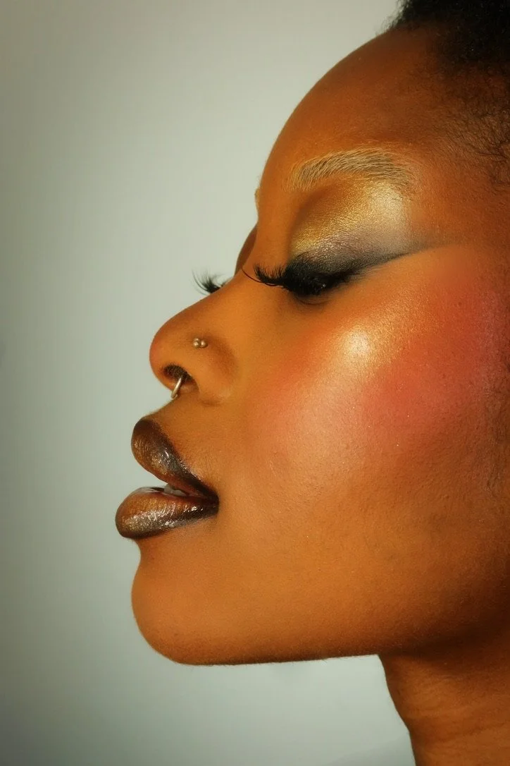 Close-up of a woman's face in profile with bold makeup, including gold eyeshadow, black eyeliner, long false eyelashes, and metallic dark lipstick. She has a nose piercing with a small hoop and a nose stud.