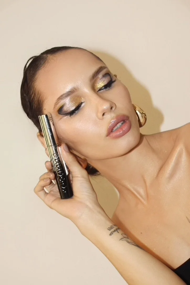 A woman with shiny makeup, gold eyeshadow, and glossy lips holding a black lipstick tube, with wet skin and a tattoo on her arm.