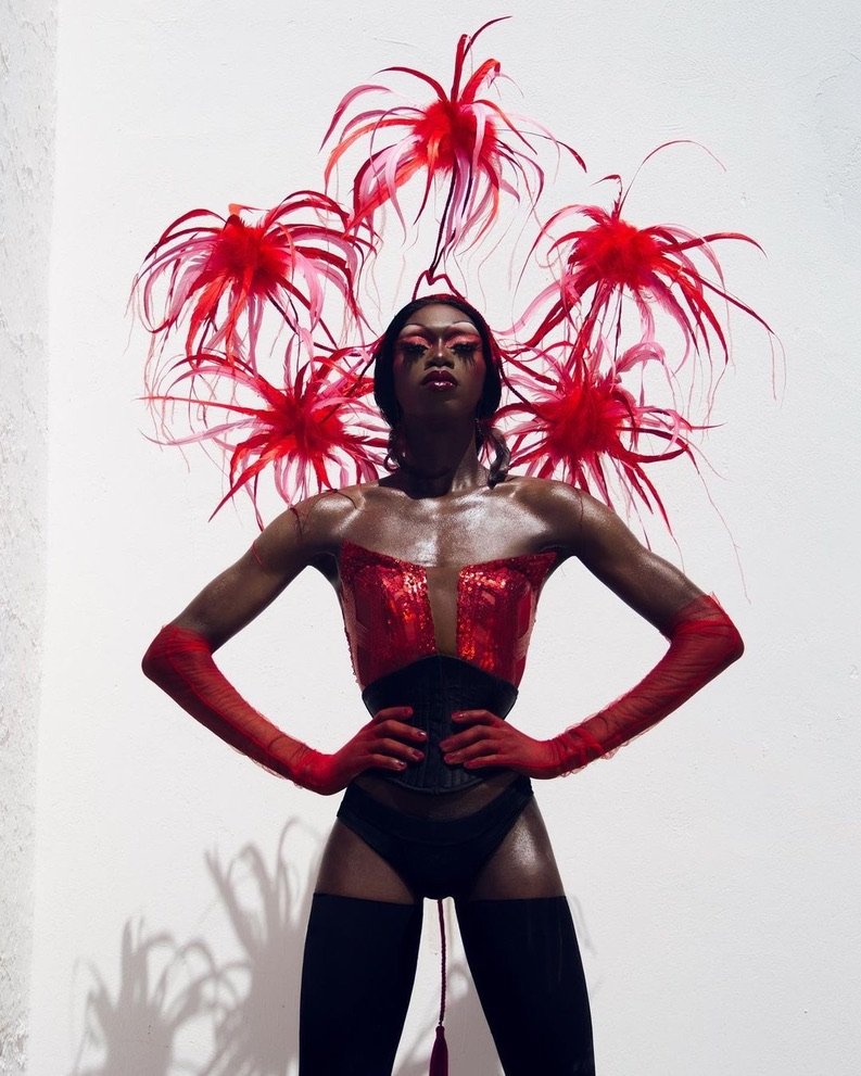 A person dressed in a flamboyant red and black costume with red feathered headpiece, standing against a white wall with hands on hips.