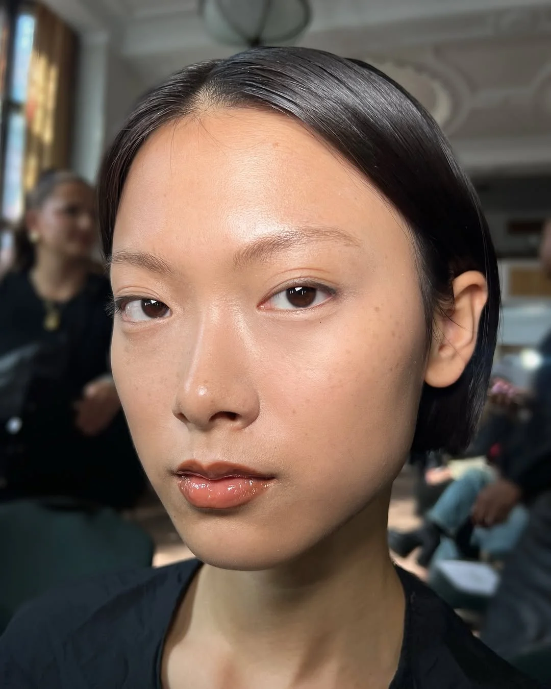 Close-up of an Asian woman with short black hair and minimal makeup in a busy indoor setting, behind her are blurred people and a bright window.