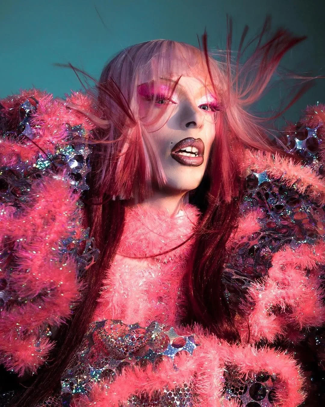 A woman with pink hair and makeup, wearing a colorful, textured outfit with feathers, sequins, and embellishments.