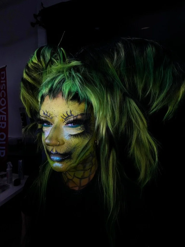 Person with green and black hair wearing spider web and makeup resembling a spider, in a dark setting.