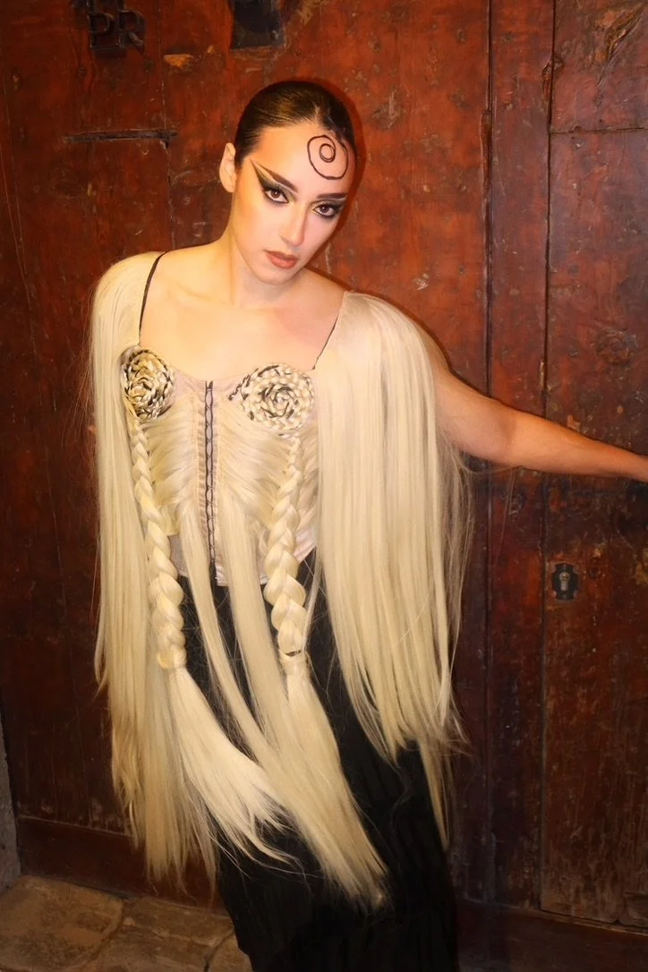 A woman with long platinum blonde hair and dramatic makeup posing in front of a rustic wooden wall.