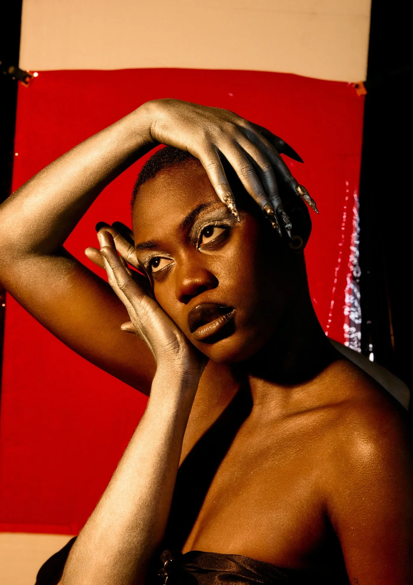 A woman with dark skin and short hair, wearing dark lipstick, posing with her hand on her forehead and looking away, in front of a red and white backdrop.