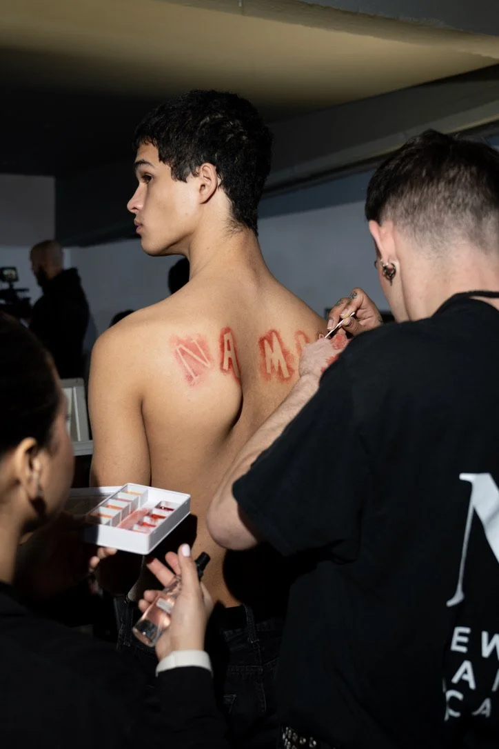 A person is getting a temporary tattoo on their back with the word 'NAMASTE' in red letters. Several people are assisting, one holding a tray with tattoo ink and others applying the tattoo.