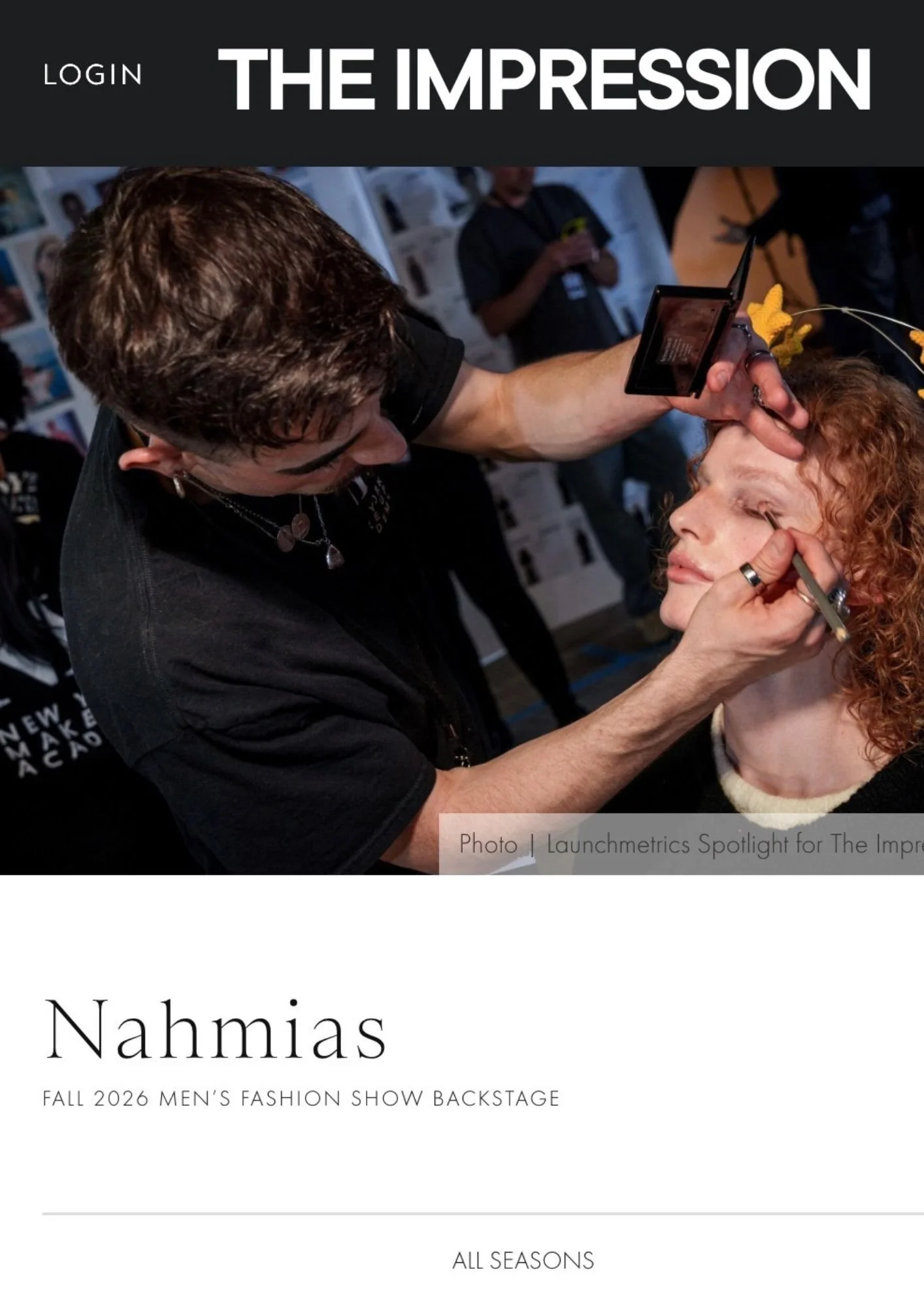 A behind-the-scenes photo of a male model backstage at a fashion show, getting makeup applied by a stylist. The setting is busy with other people in the background, and there is a wall with numerous pictures and posters.