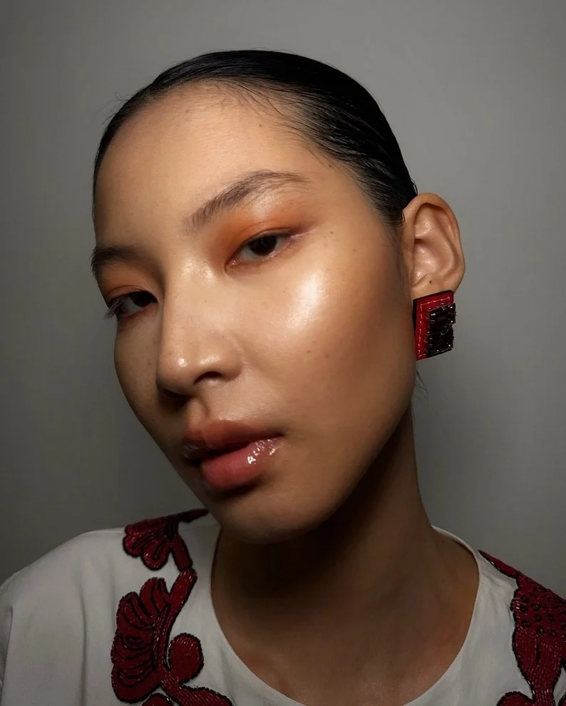 A woman with dark hair slicked back, wearing makeup with warm tones, glossy lips, and large red and black earrings, posing against a gray background.