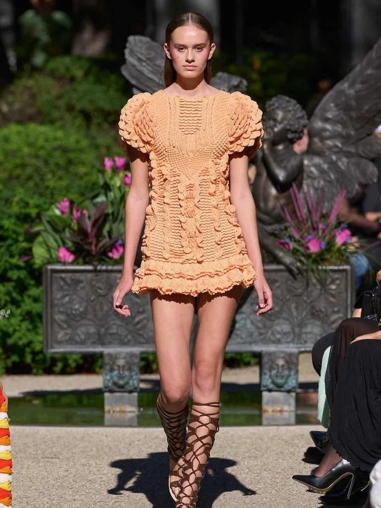 Model walking down the runway wearing an orange textured dress with puffed sleeves, paired with brown gladiator-style sandals. The background features plants, flowers, and a decorative sculpture.