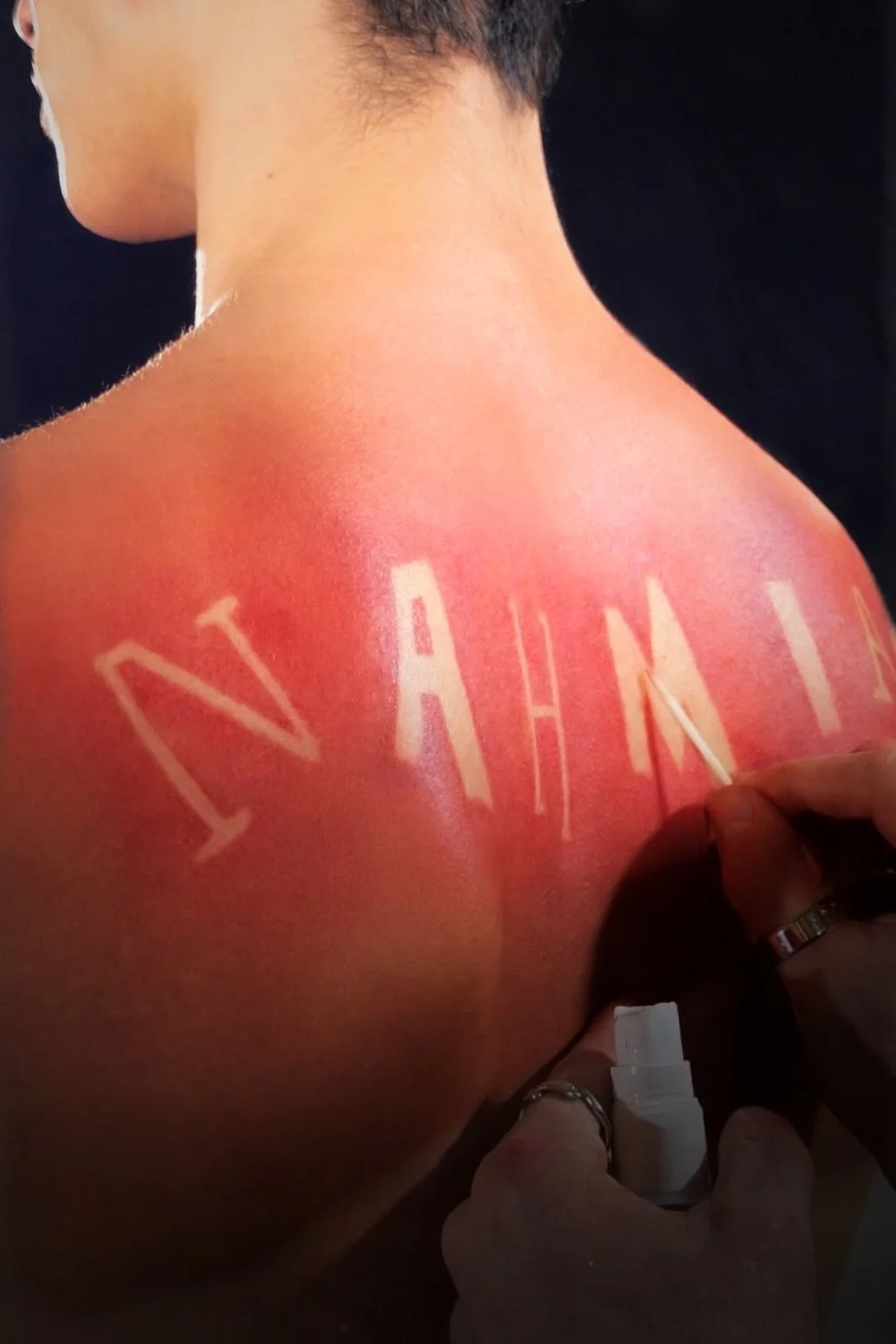 A person with short dark hair is showing their back, which has the word 'VANITY' being painted on with white paint.