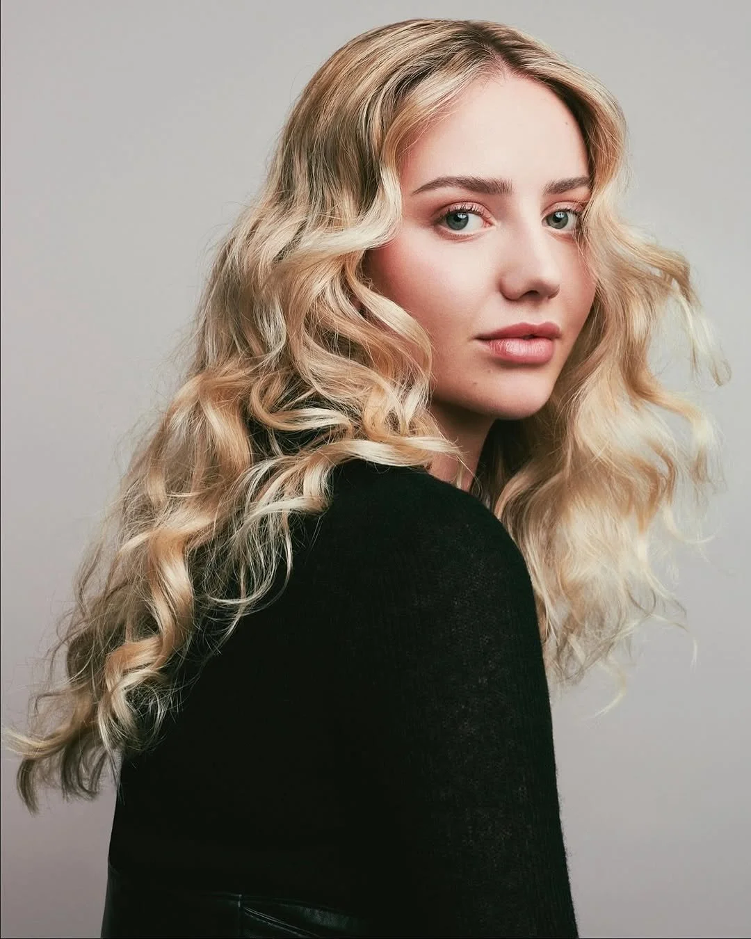 Portrait of a young woman with long, curly blonde hair wearing a black top, looking at the camera with a neutral expression against a plain, light gray background.