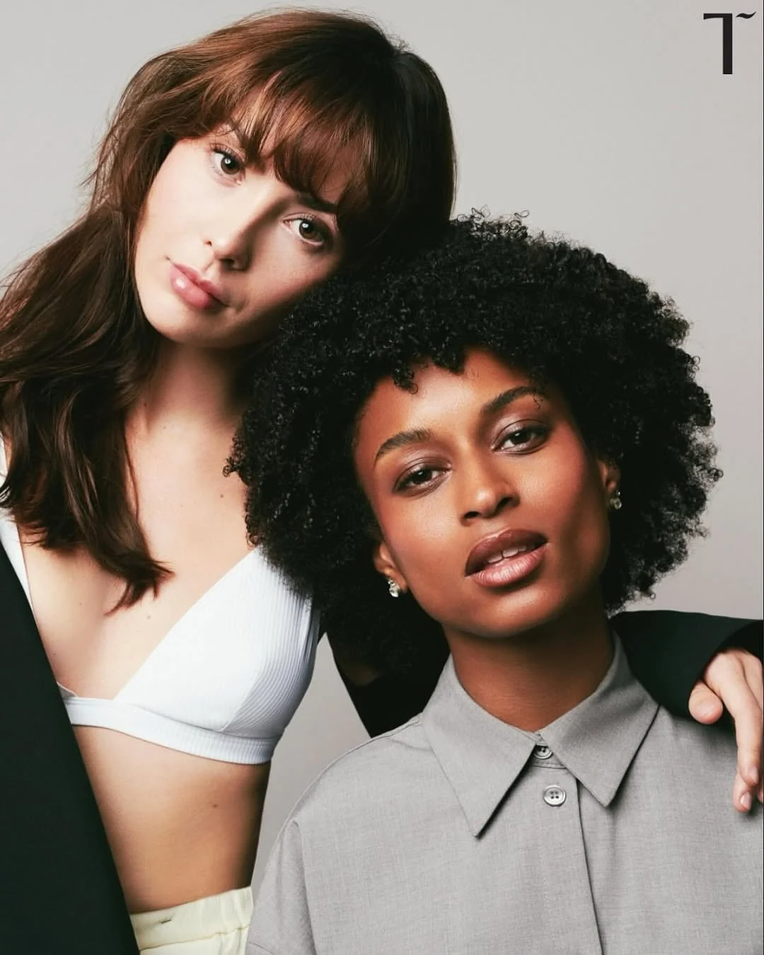 Two women with contrasting hair styles, one with straight brown hair and the other with curly black hair, posing together against a plain background.
