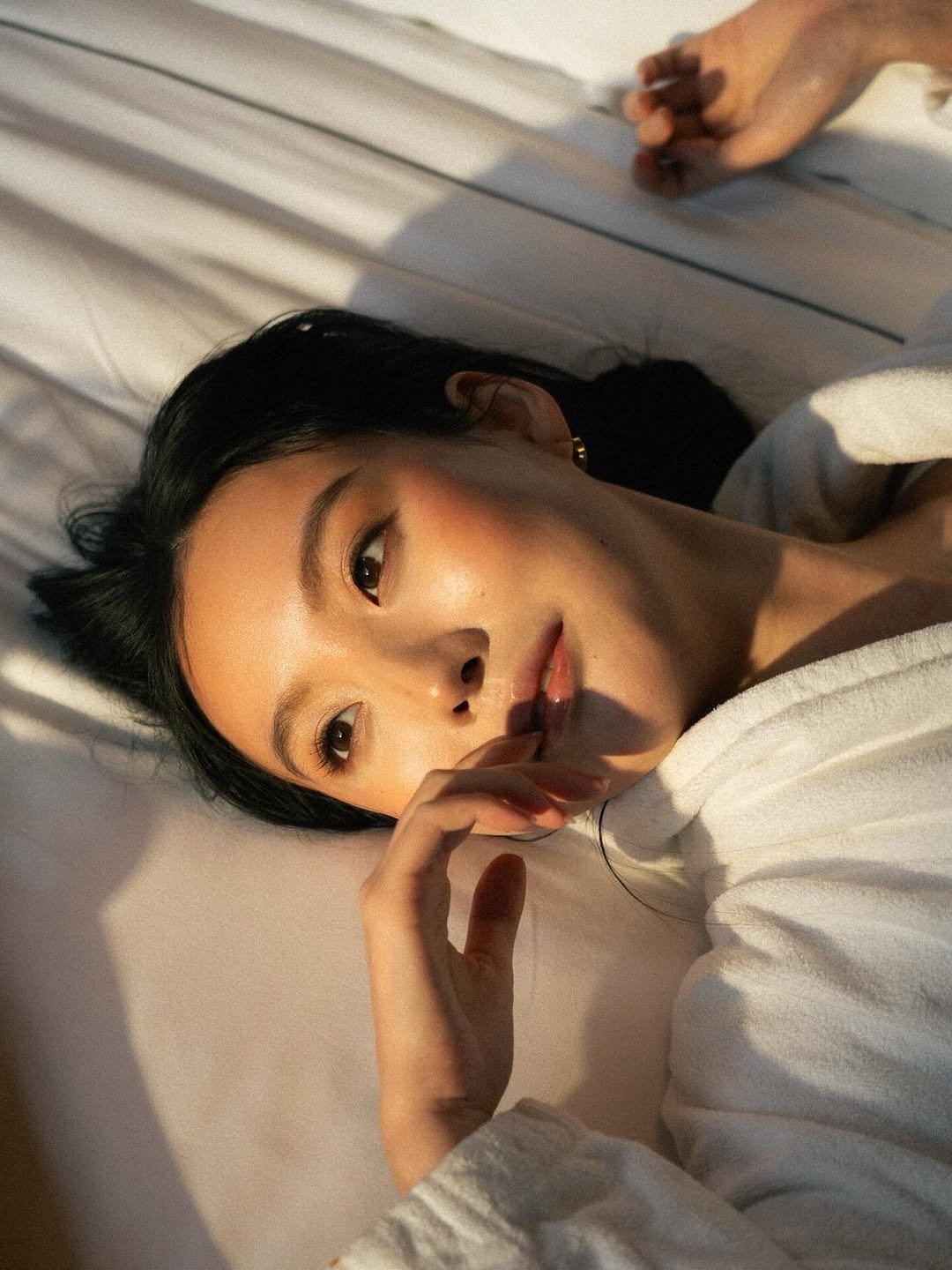 A young woman with black hair lying on her side on a pillow, looking into the camera with her hand near her face, in a softly lit setting.