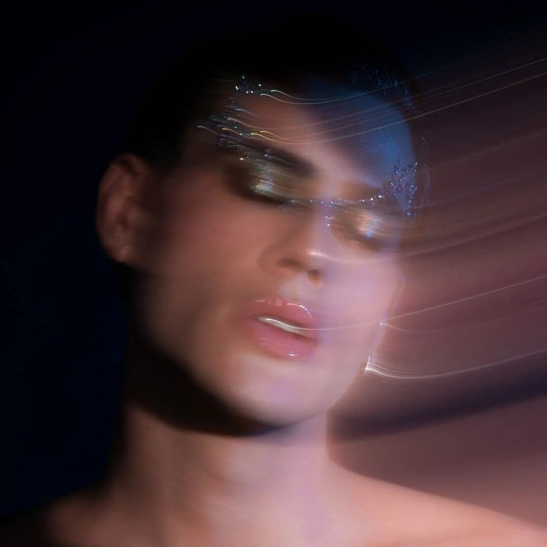 A woman with makeup, wearing glasses, appears to have motion blur or light trails across her face, creating an abstract, artistic effect.