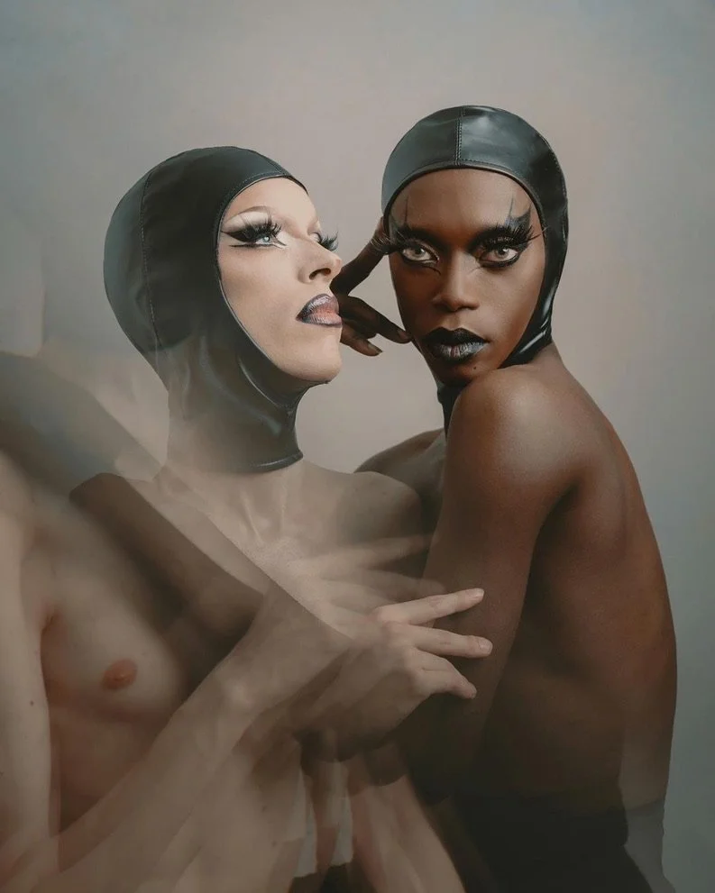 Two topless women wearing black leather head coverings with makeup and striking eye looks, posing closely together against a neutral background.