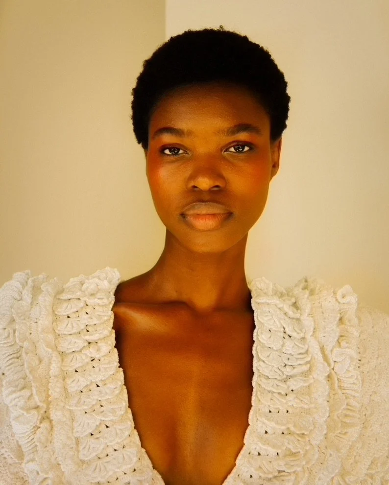 Portrait of a woman with short curly hair wearing a textured white dress with ruffle details, standing against a plain background.