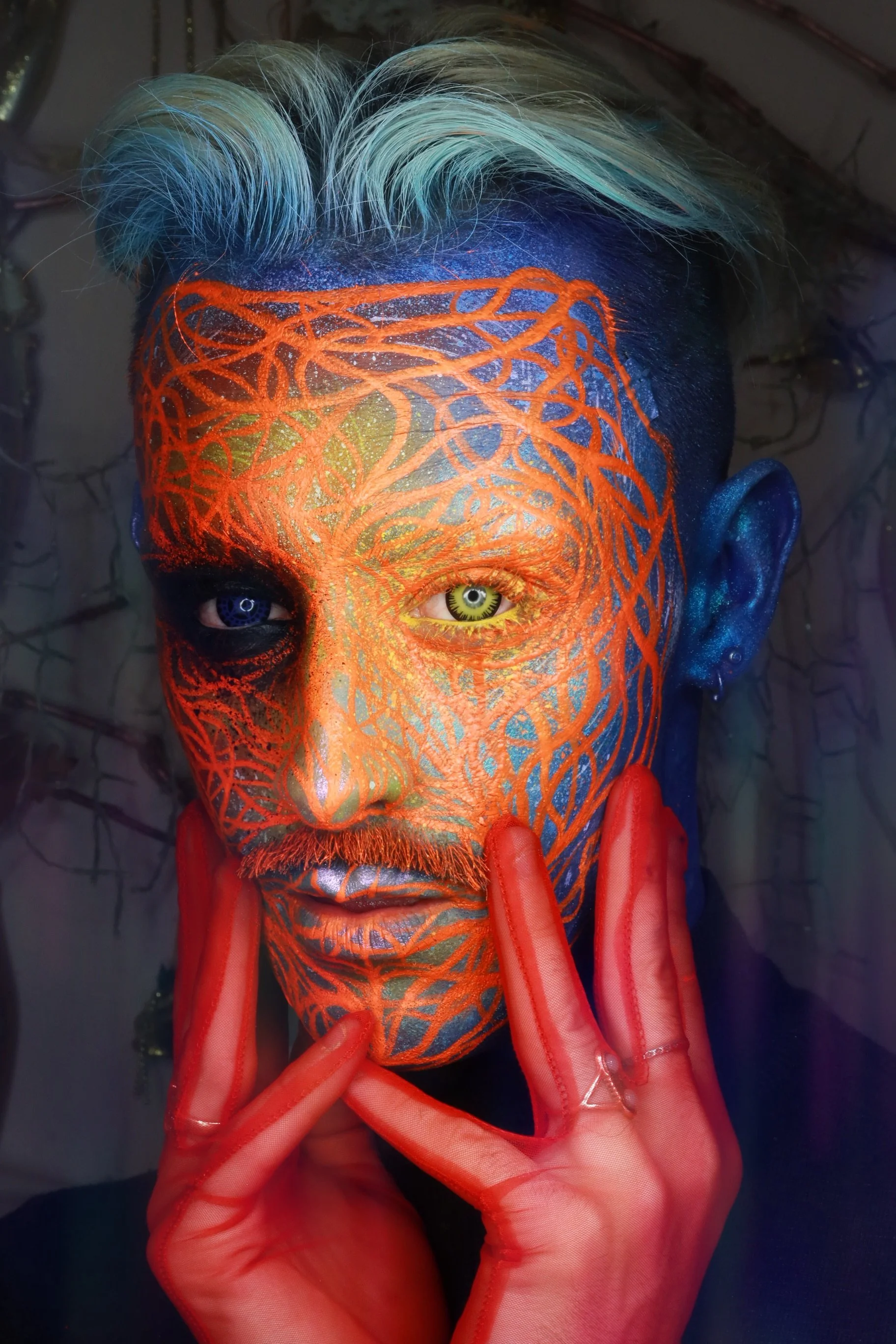A person with short, multi-colored hair and face painted with an orange, intricate web-like pattern, holding their face with red, translucent gloves.
