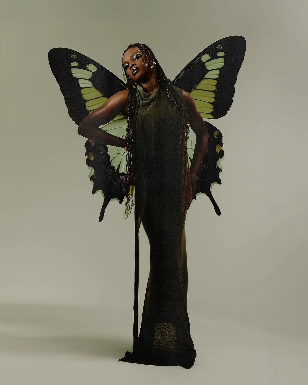 Woman dressed as a butterfly with black and yellow wings, dark green dress, standing with hands on hips against a plain background.