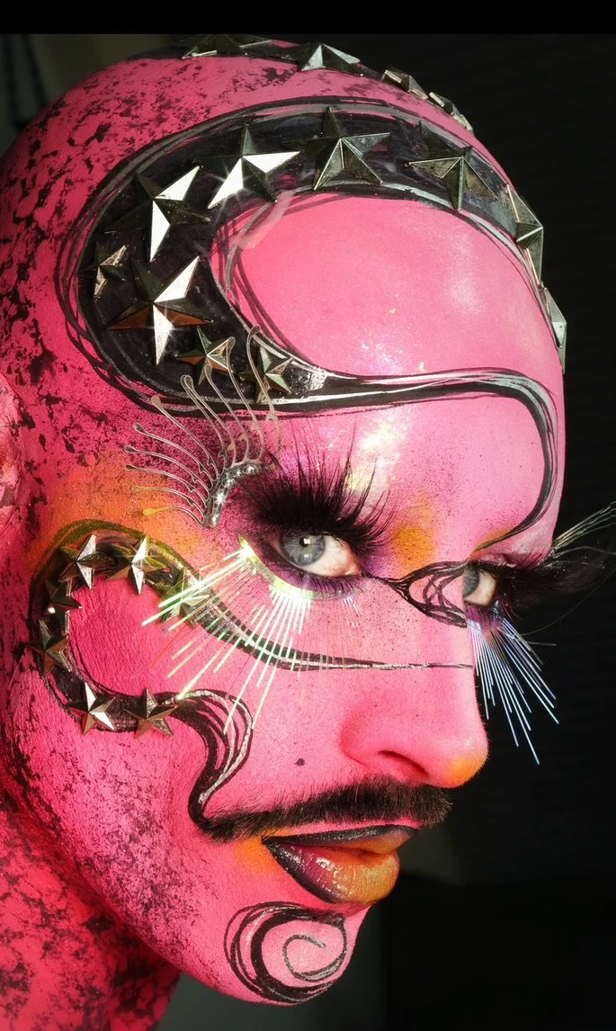 Close-up of a person with vibrant pink face paint, metallic star-shaped accessories around the eye and forehead, artistic black and silver painted designs, elaborate false eyelashes, and colorful makeup.
