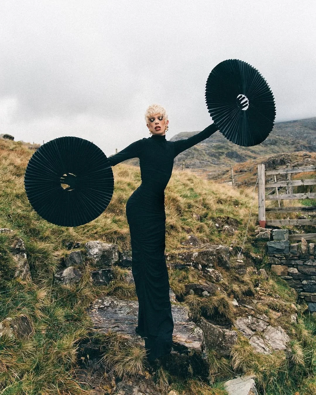 A woman with platinum blonde hair is standing outdoors on rocky and grassy terrain, wearing a fitted long black dress and holding two large black umbrellas.
