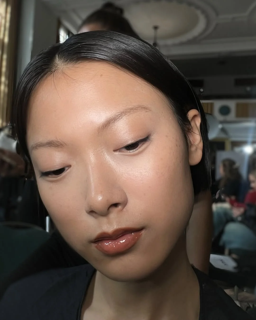 Close-up of a young woman's face with short black hair, natural makeup, and glossy lips in an indoor setting with people and a ceiling light in the background.