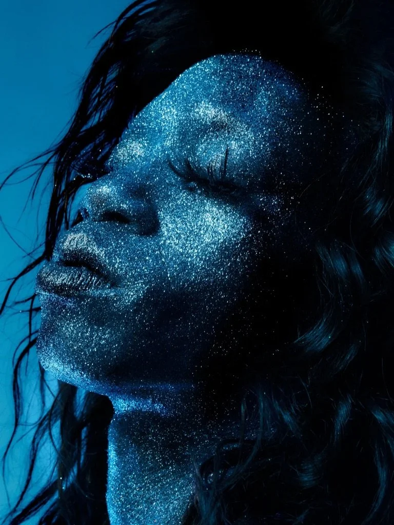 A person with long dark hair has their face and neck covered in blue glitter, with their eyes closed and head slightly tilted back against a blue background.