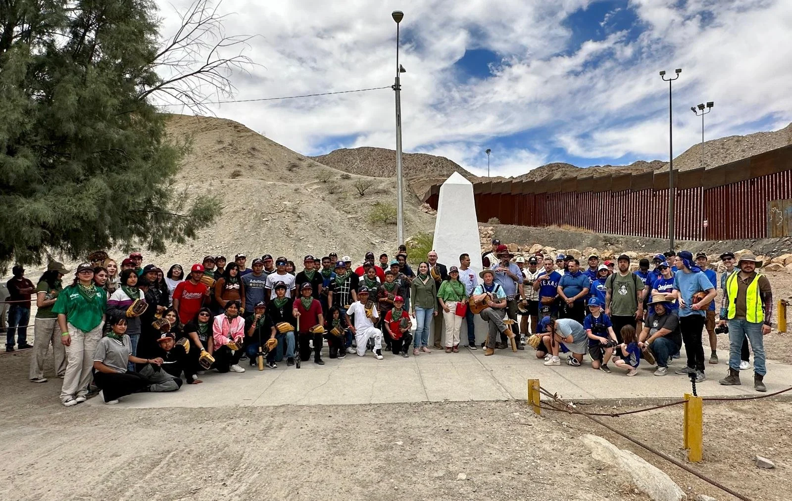 Group of people gathered outdoors in a desert-like area with mountains in the background, some wearing baseball gloves and holding sports equipment, standing in front of a white monument and a tall border wall.