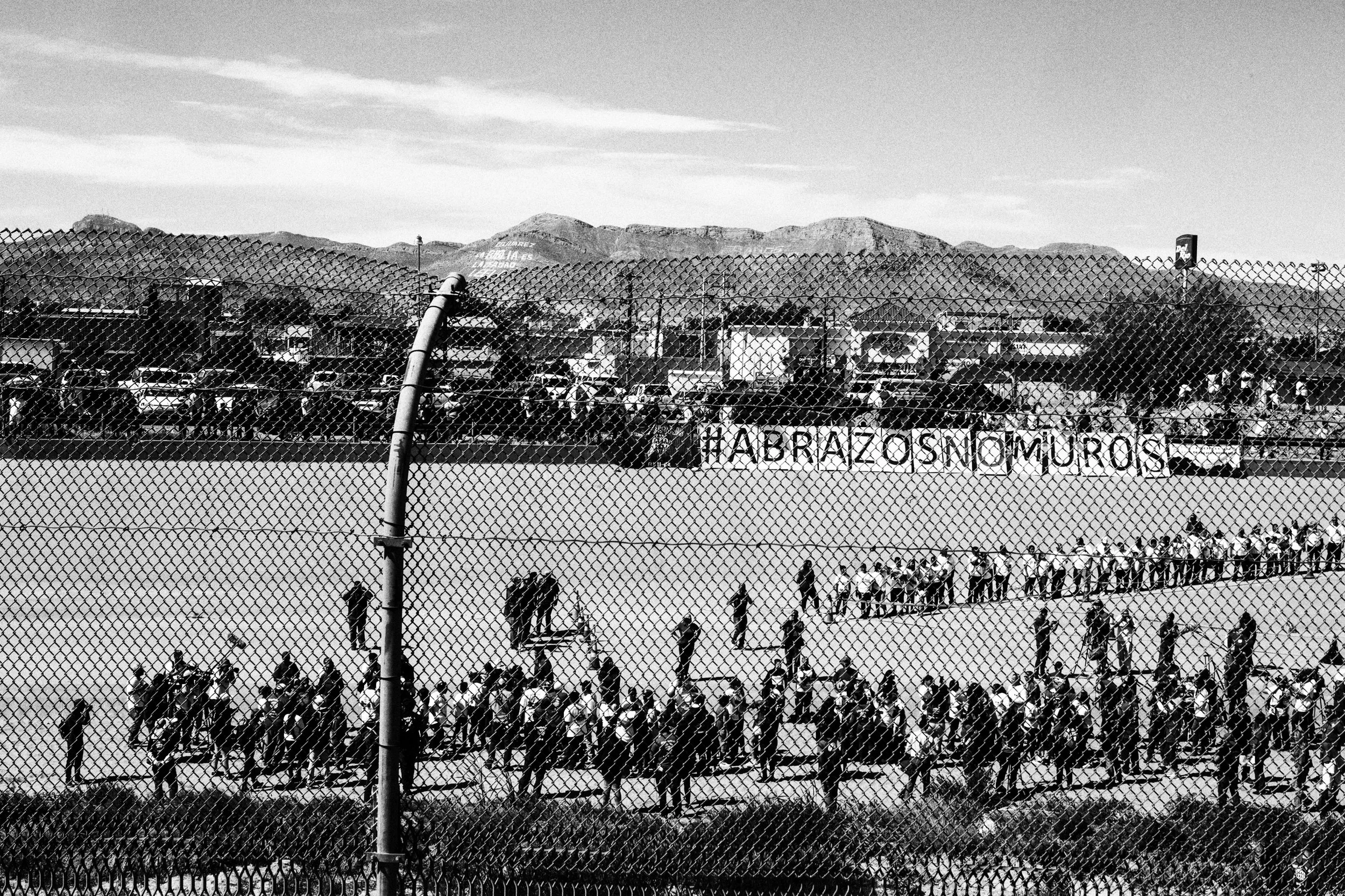 Black and white photo of people standing in a line behind a chain-link fence, with mountains in the background and a sign that appears to say 'FABRICA OS MOURO.'