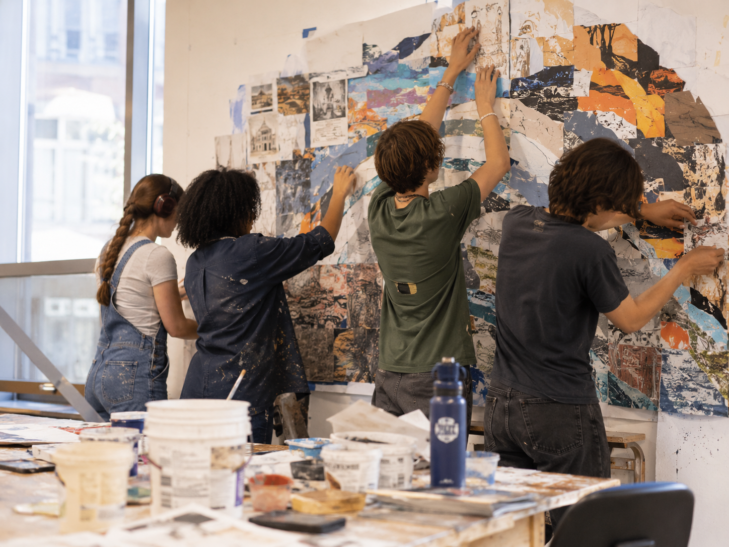 Group of five people working on a collage mural in an art studio, with art supplies on a table in the foreground, large window in the background.
