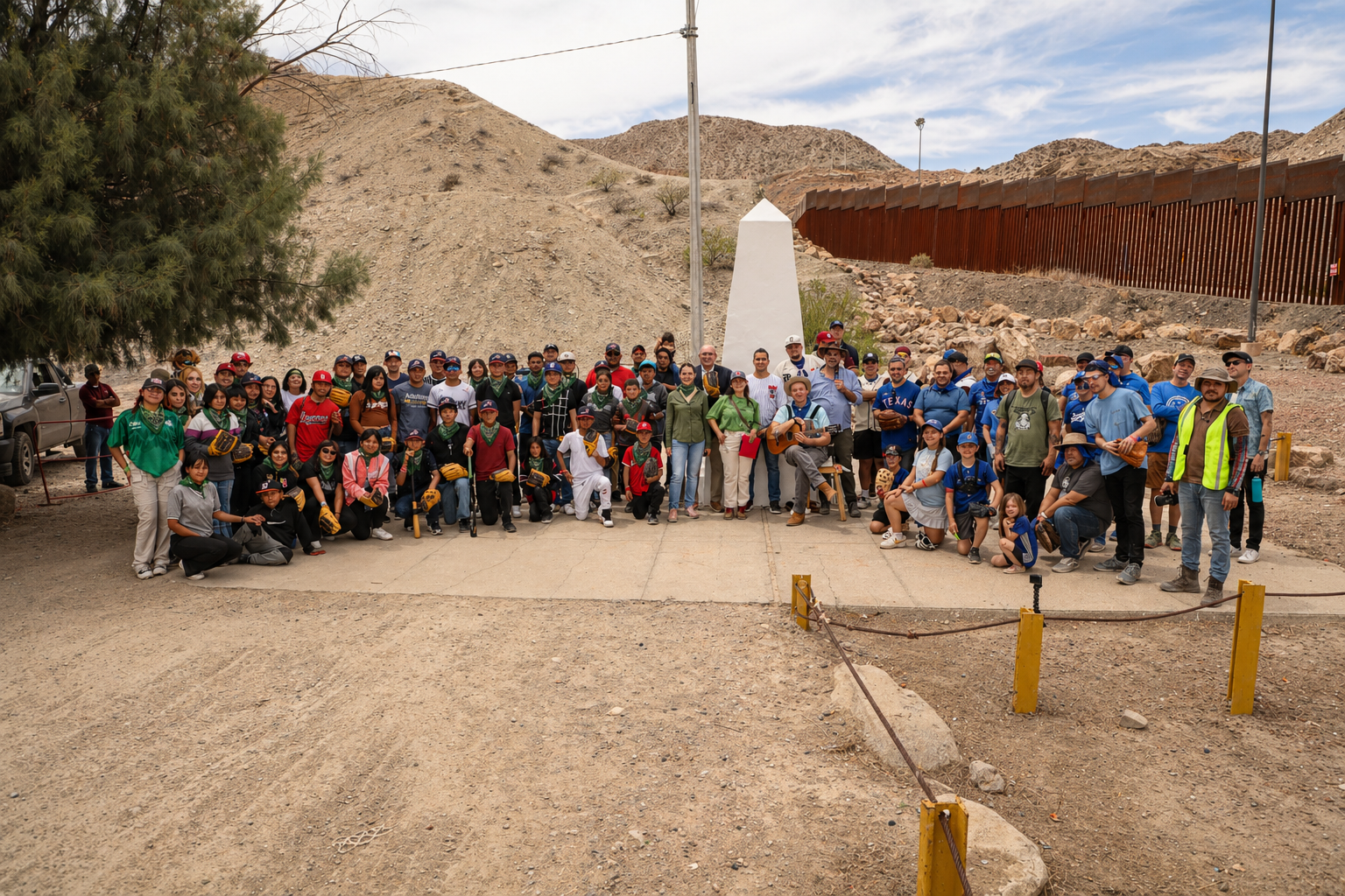 Group of people gathered outdoors in a desert-like area with mountains in the background, some wearing baseball gloves and holding sports equipment, standing in front of a white monument and a tall border wall.